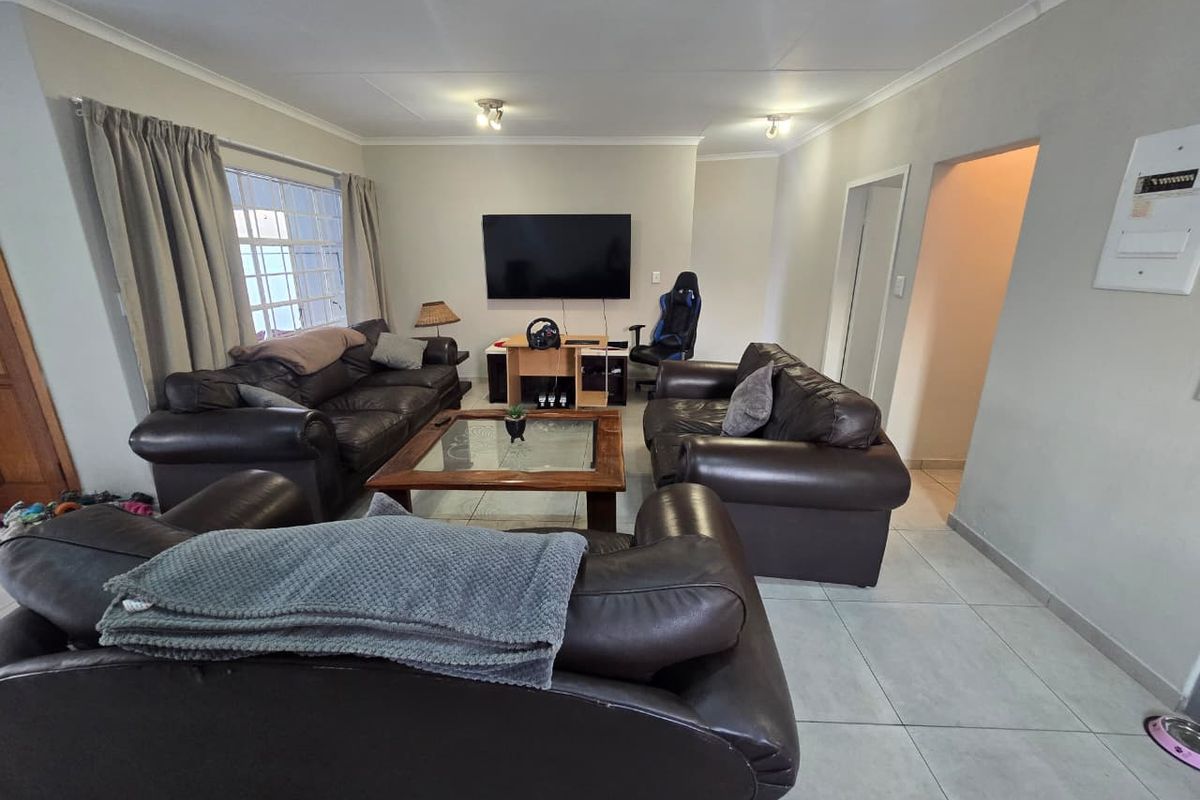 A spacious living room featuring neutral tiled flooring, ceiling-mounted lighting, and a large window with security bars, offering a comfortable and functional layout.