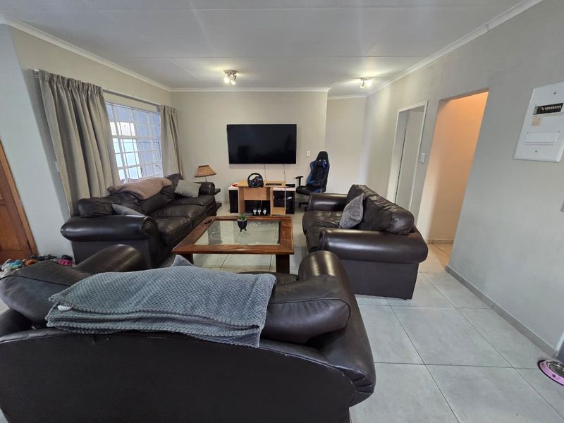 A spacious living room featuring neutral tiled flooring, ceiling-mounted lighting, and a large window with security bars, offering a comfortable and functional layout.