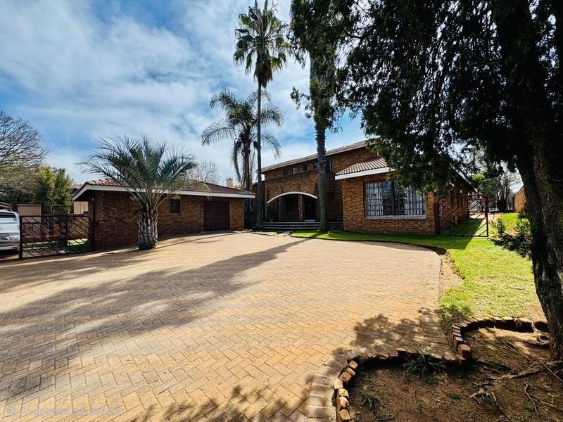 A spacious front exterior view of a brick-faced house featuring a large paved driveway, attached garage, and established garden surroundings.