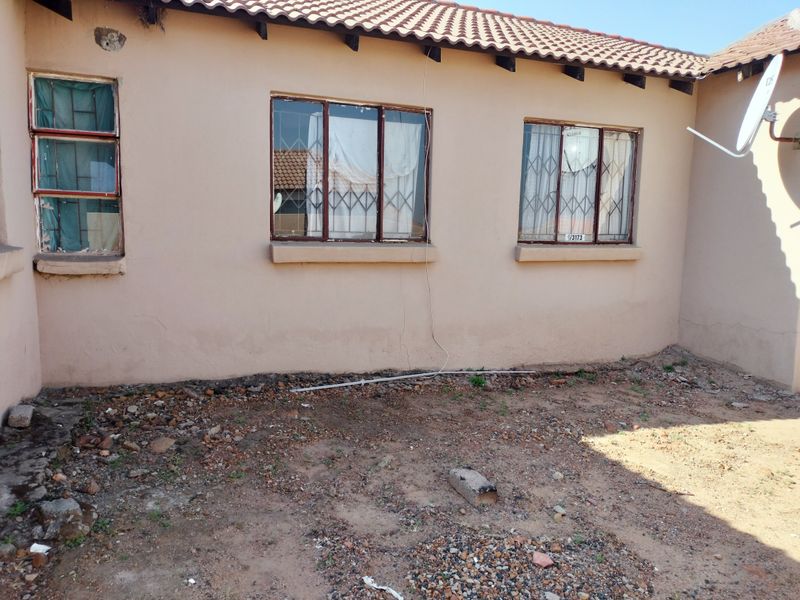 Rear exterior view of the house showing tiled roofing, security bars on windows, and an unlandscaped yard area requiring maintenance.