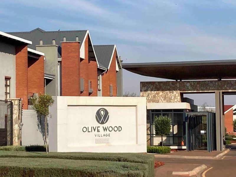 The entrance to Olive Wood Village, featuring a modern security gatehouse, stone-clad architectural elements, and well-maintained brick townhouse facades.