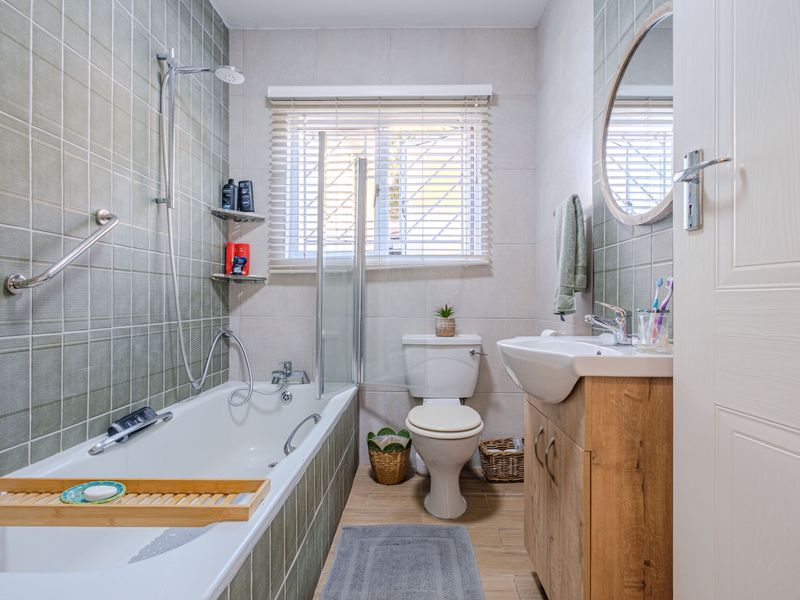 A well-maintained bathroom featuring a bathtub with shower attachment, a modern vanity unit, and neutral wall tiling with a window for natural light.
