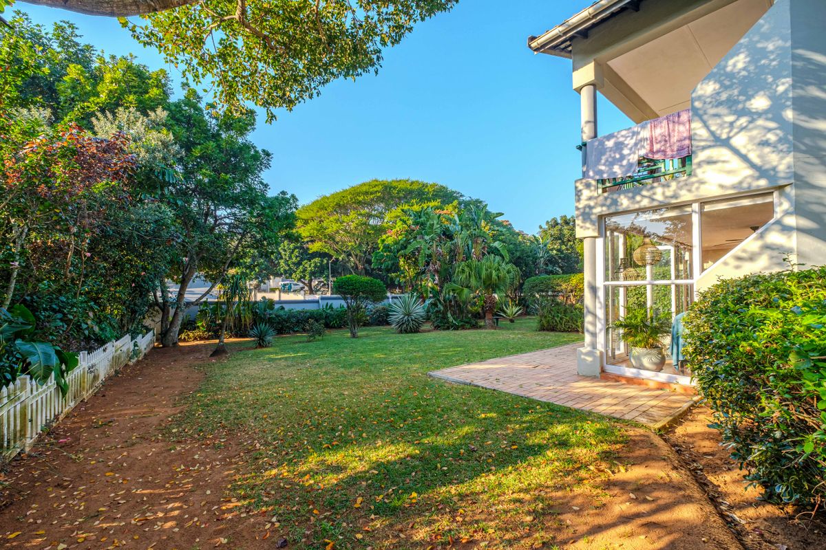 A spacious rear garden featuring a paved patio area, a balcony above, and a well-maintained lawn surrounded by mature trees and a picket fence.