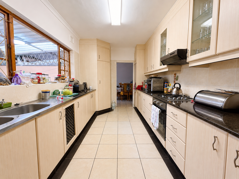 A functional galley-style kitchen featuring light wood cabinetry, granite countertops, a gas hob, and tiled flooring with ample storage space.