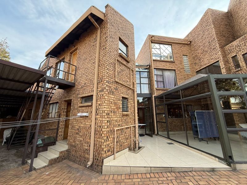 Rear exterior view of a multi-story face brick house featuring a metal staircase, paved patio area, and a glass-enclosed conservatory structure.
