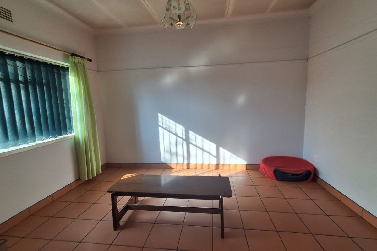 A spacious, sunlit living room featuring neutral walls, tiled flooring, and a large window with vertical blinds.