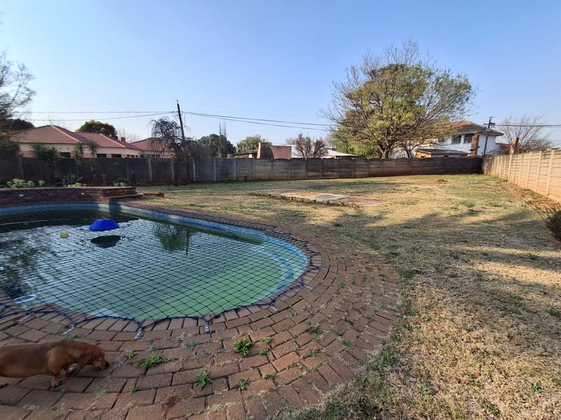 Spacious backyard featuring a swimming pool with a safety net, brick paving, and a perimeter wall, set within a large grassed garden area.