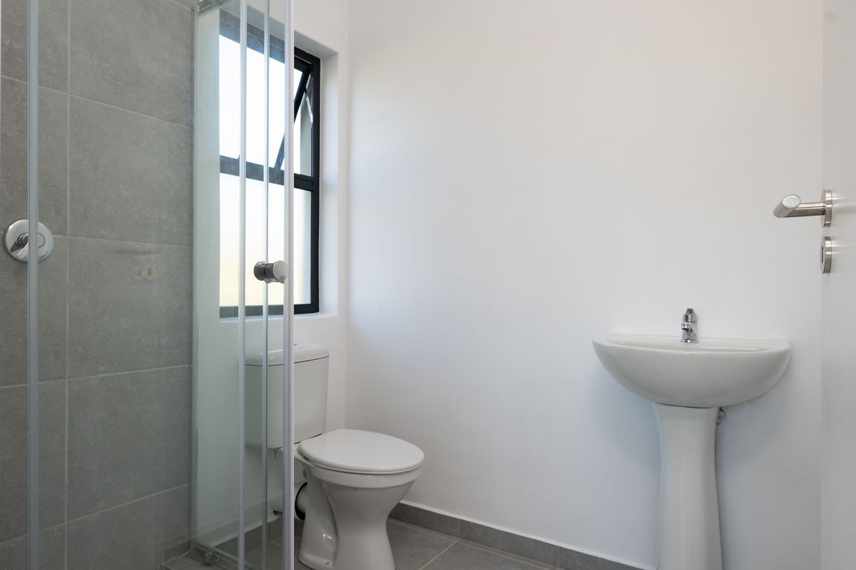 A clean, modern bathroom featuring a pedestal basin, toilet, and a glass-enclosed shower with grey tiled accents and a top-hung window.