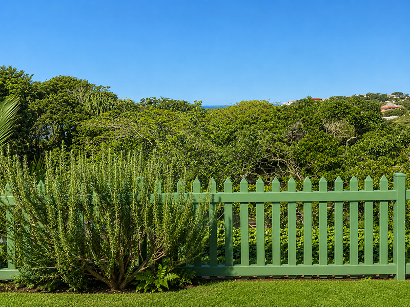 A scenic view of lush greenery and distant coastal horizon, framed by a well-maintained green picket fence.