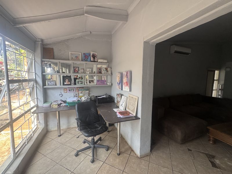 A functional home office space featuring tiled flooring, built-in shelving, and security bars on the windows, adjacent to a living area with air conditioning.
