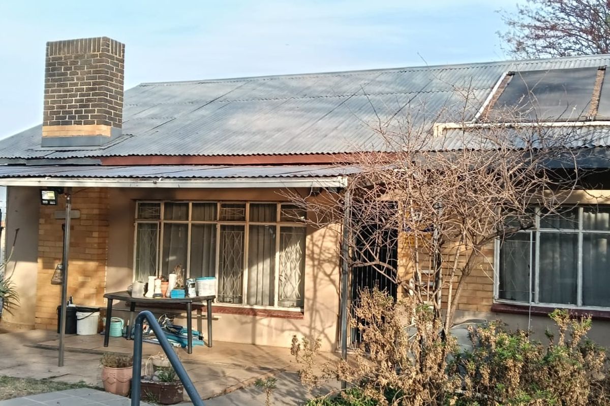 Rear view of a single-story house featuring a corrugated iron roof, brick chimney, solar water heating panels, and a covered patio area.