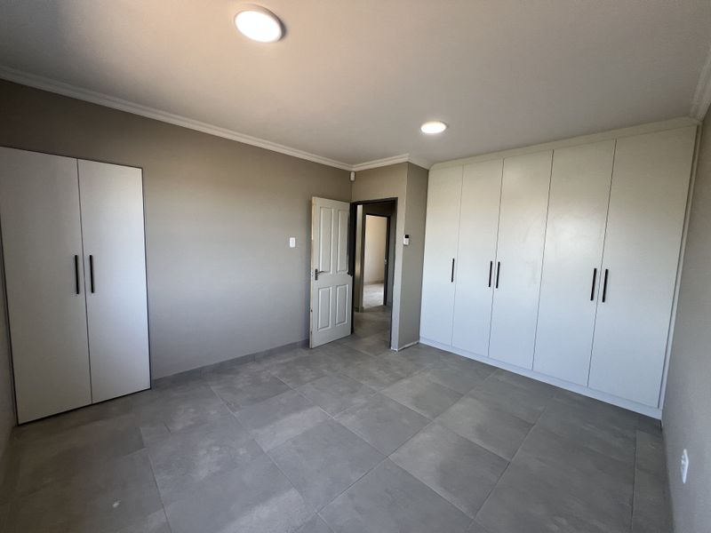 A spacious, empty bedroom featuring neutral tiled flooring, ample built-in cupboard storage, and modern recessed ceiling lighting.