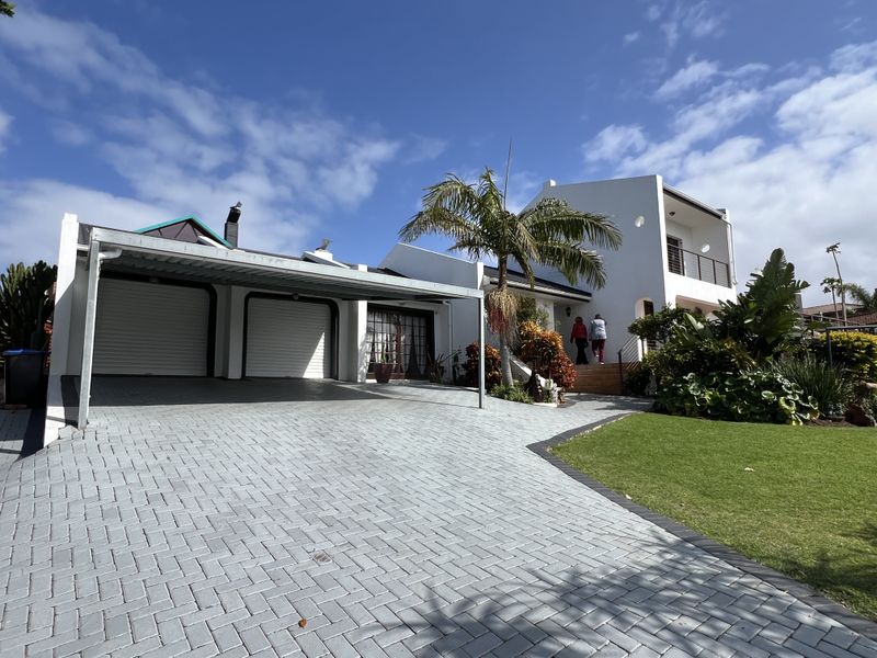 Front view of a well-maintained house featuring a paved driveway, double garage, metal carport, and a landscaped garden with a balcony.
