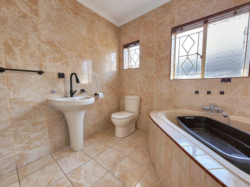 A functional bathroom featuring a pedestal basin, toilet, and a built-in bathtub with full-height wall tiling and security bars on the windows.