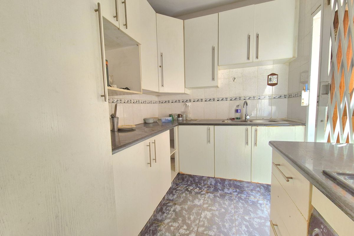 A compact kitchen featuring white built-in cabinetry, laminate countertops, a stainless steel sink, and tiled flooring with a security gate at the entrance.