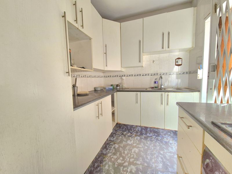 A compact kitchen featuring white built-in cabinetry, laminate countertops, a stainless steel sink, and tiled flooring with a security gate at the entrance.