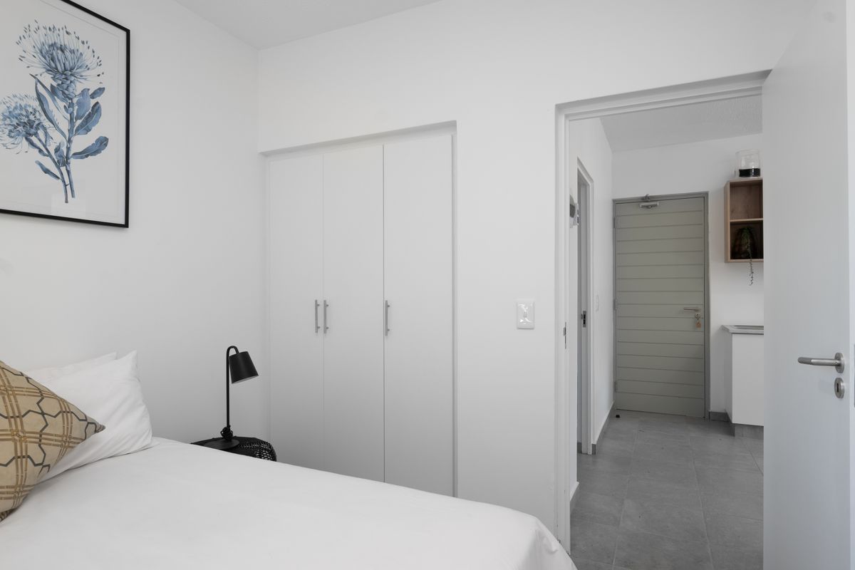 A clean, modern bedroom featuring built-in wardrobes and tiled flooring, with a view into the adjacent hallway and entrance area.