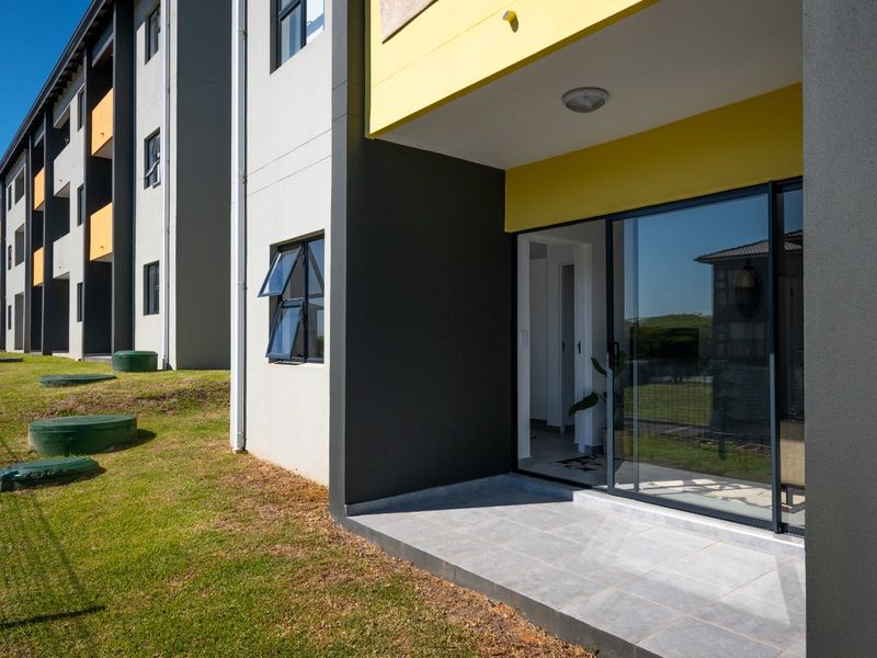 A ground floor apartment patio featuring tiled flooring and a sliding glass door entrance, set against a modern building facade with landscaped lawn.