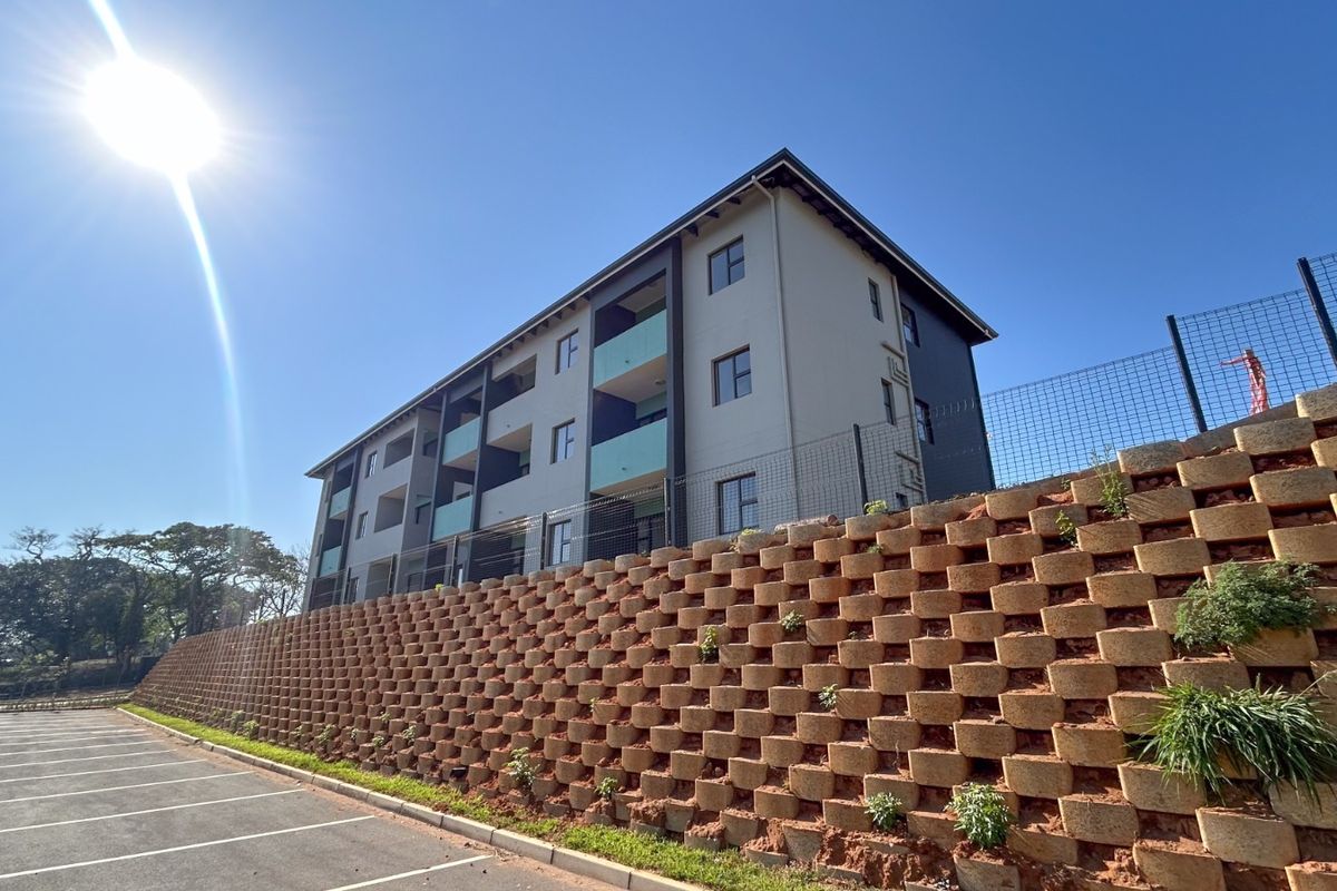 Exterior view of a modern apartment building featuring balconies, secure perimeter fencing, and a large tiered retaining wall overlooking a paved parking area.