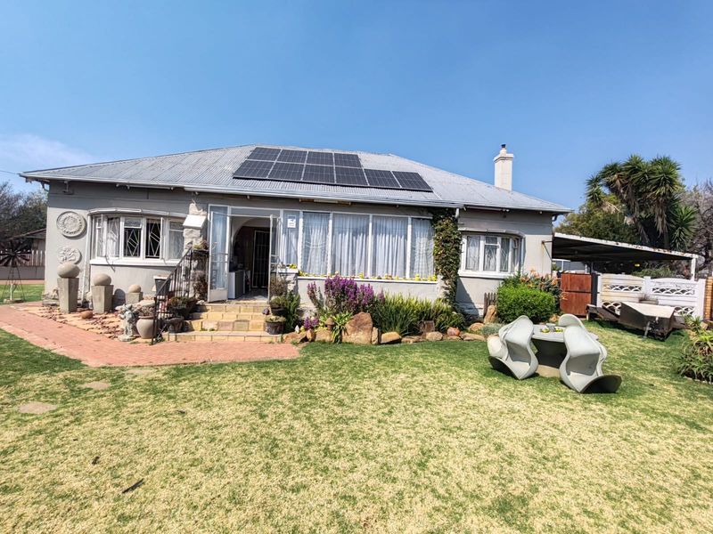 Rear view of a single-story house featuring a corrugated metal roof with solar panels, a brick paved walkway, and a well-established garden.