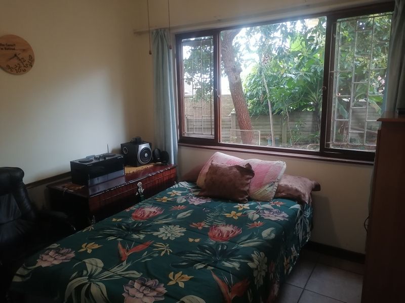 A bedroom featuring tiled flooring and a large window with security bars, offering a view of the garden.