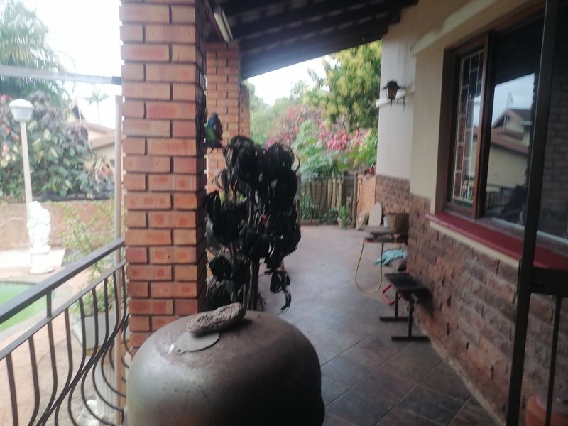 A covered patio area featuring exposed brick columns, tiled flooring, and wrought iron railings, overlooking the garden and pool area.