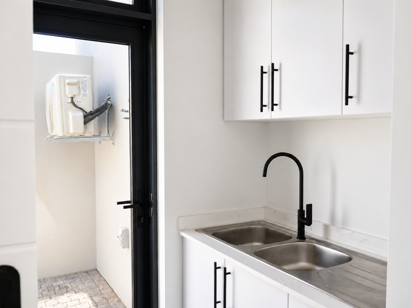 A clean, modern scullery featuring white cabinetry, stone countertops, a double stainless steel sink with a black mixer tap, and direct access to an outdoor area.