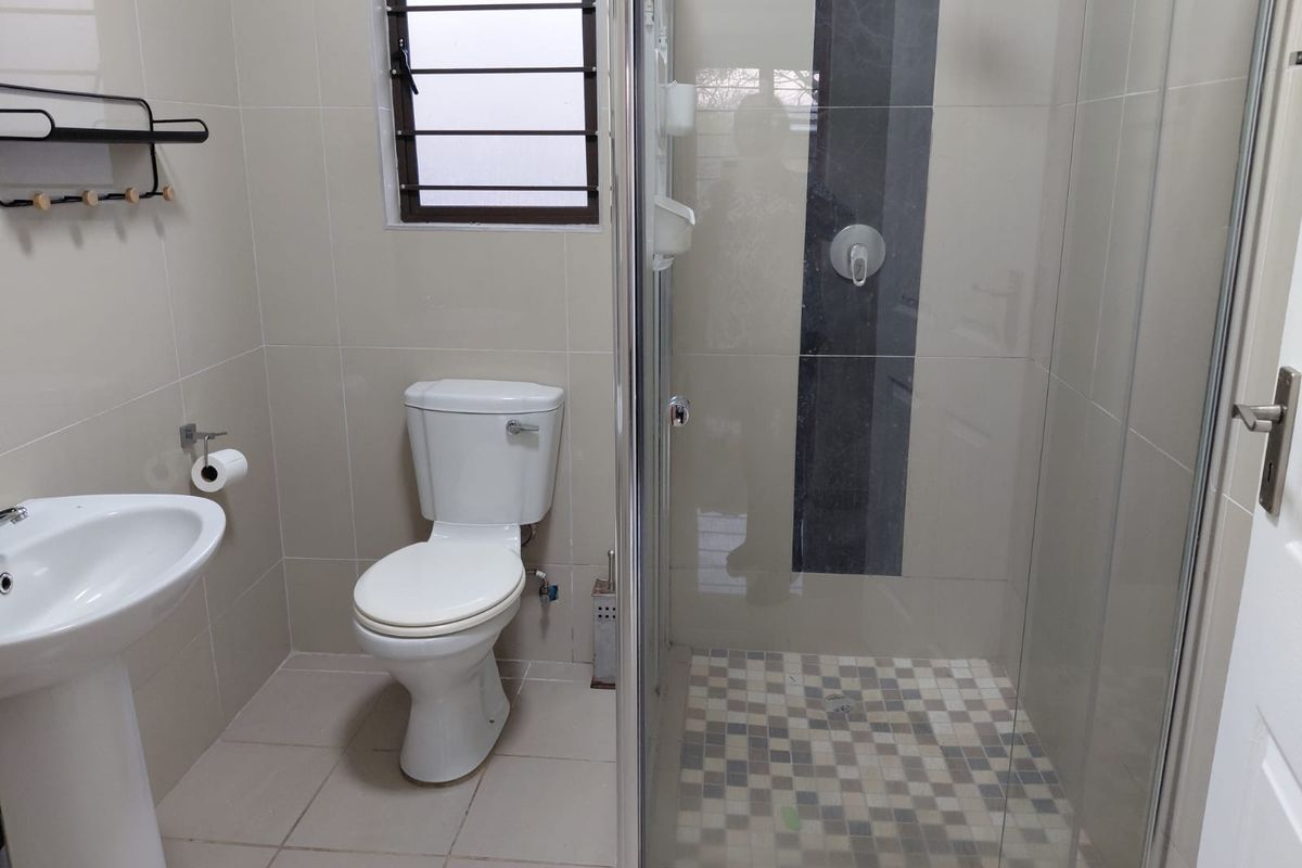 A clean, modern bathroom featuring a pedestal basin, toilet, and a glass-enclosed shower with mosaic floor tiling and neutral wall tiles.
