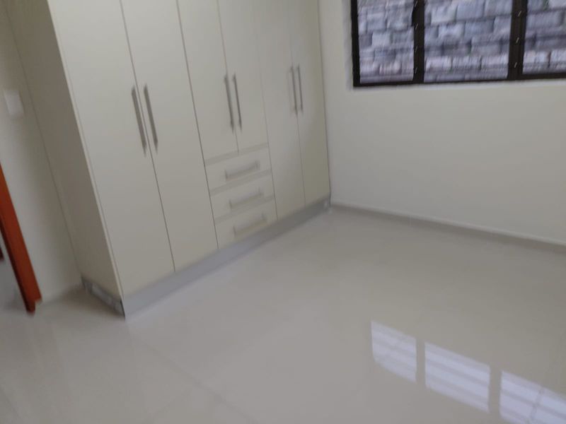 A clean, empty bedroom featuring neutral tiled flooring and ample built-in cupboard storage with a window overlooking an exterior brick wall.