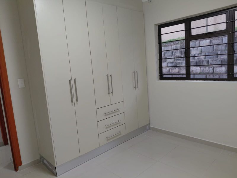 A clean, empty bedroom featuring modern built-in cupboards, neutral tiled flooring, and a window fitted with security bars.