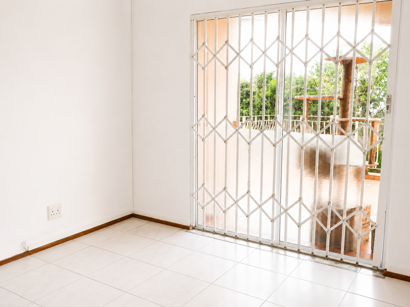 A bright, empty living area featuring neutral tiled flooring and a sliding glass door with a security gate leading to an outdoor patio.