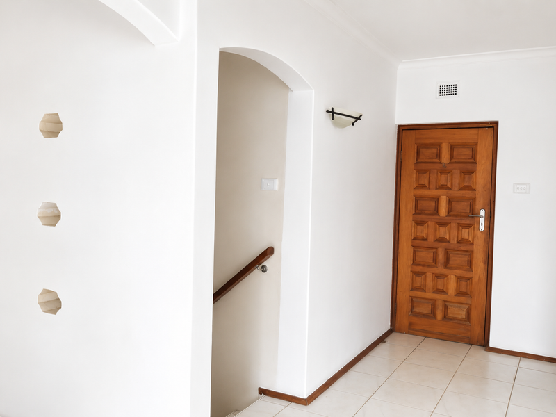 A bright, empty hallway featuring ceramic floor tiles, an arched entryway leading to a staircase, and a classic wooden panelled door.
