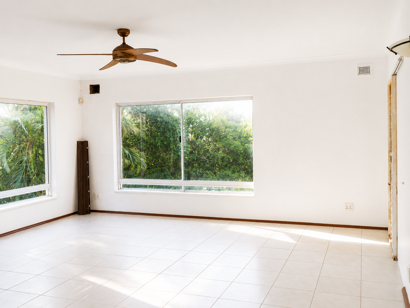 A bright, empty living room featuring tiled flooring, a ceiling fan, and large windows overlooking greenery, with an unfinished doorway awaiting final trim.