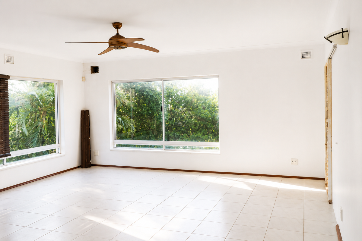 A bright, empty living room featuring tiled flooring, a ceiling fan, and large windows overlooking greenery, with an unfinished doorway awaiting final trim.