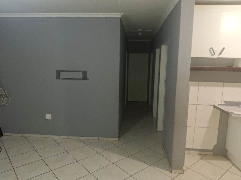 An open-plan living area featuring tiled flooring, a hallway leading to internal rooms, and a partially visible kitchen with built-in cabinetry.