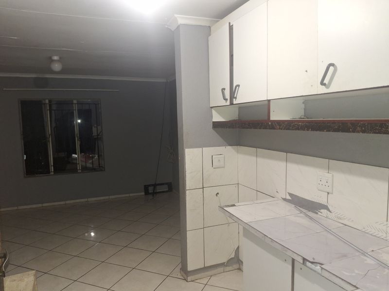 Open plan living area featuring tiled flooring and a kitchen space with built-in cabinetry, requiring some cosmetic repairs and updates.