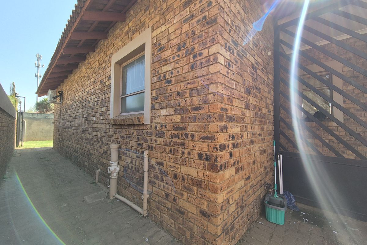Side view of the property featuring exposed brick exterior, tiled roof, and paved walkway with external plumbing and security gate access.