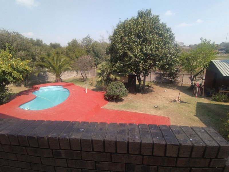 An elevated view of a backyard featuring a swimming pool with a red paved surround, mature trees, and a brick boundary wall.