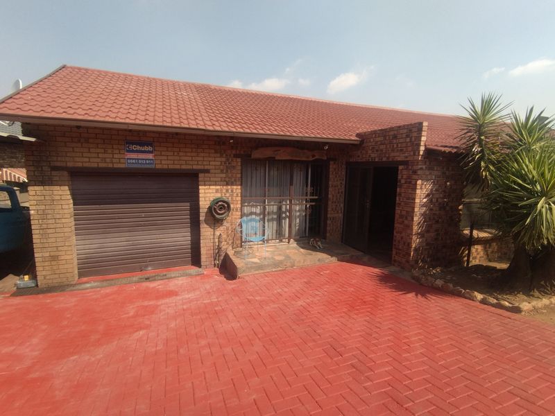 Front elevation of a brick townhouse featuring a single roller garage door, paved driveway, and a covered entrance area with security gate.