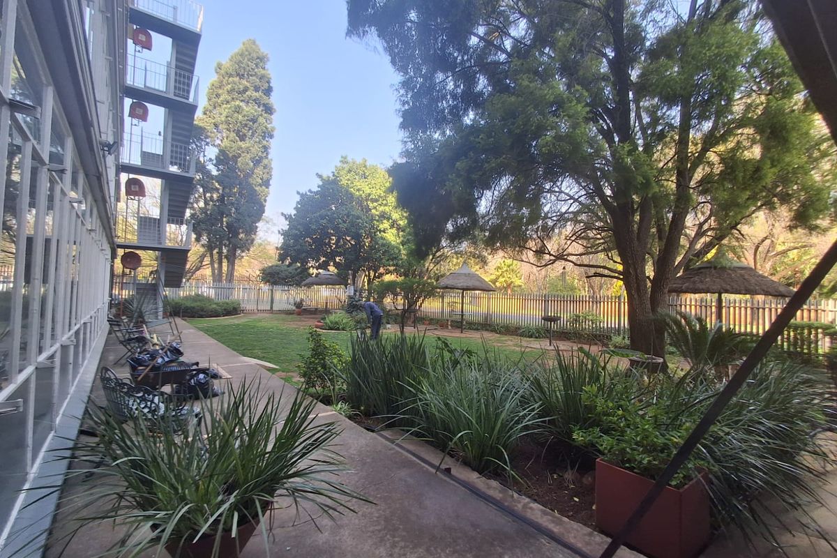 A well-maintained rear garden area featuring a paved walkway, mature trees, landscaped greenery, and a metal fire escape staircase attached to the building.