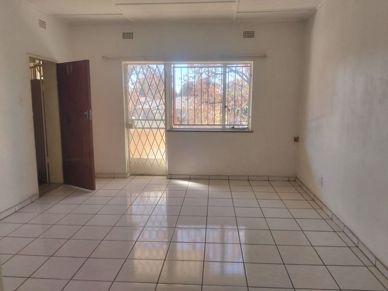A bright, empty living area featuring neutral tiled flooring, a security gate, and burglar bars on the window.