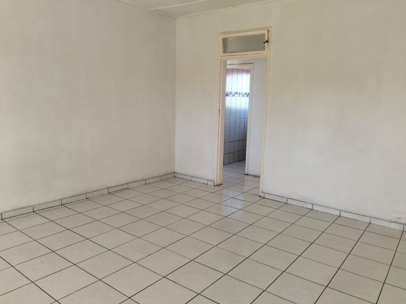 An empty living area featuring white walls and neutral ceramic floor tiles, leading through a doorway into an adjacent tiled space.
