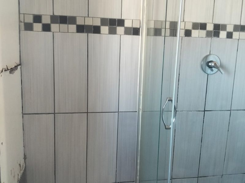 A bathroom shower area featuring tiled walls and flooring with a glass shower enclosure, showing visible signs of wear and wall damage.