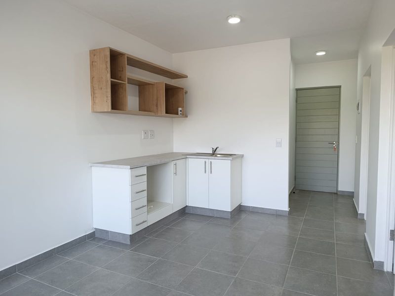 A clean, modern kitchen area featuring white cabinetry, wood-look shelving, grey tiled flooring, and a stainless steel sink, presented in a vacant, well-maintained state.