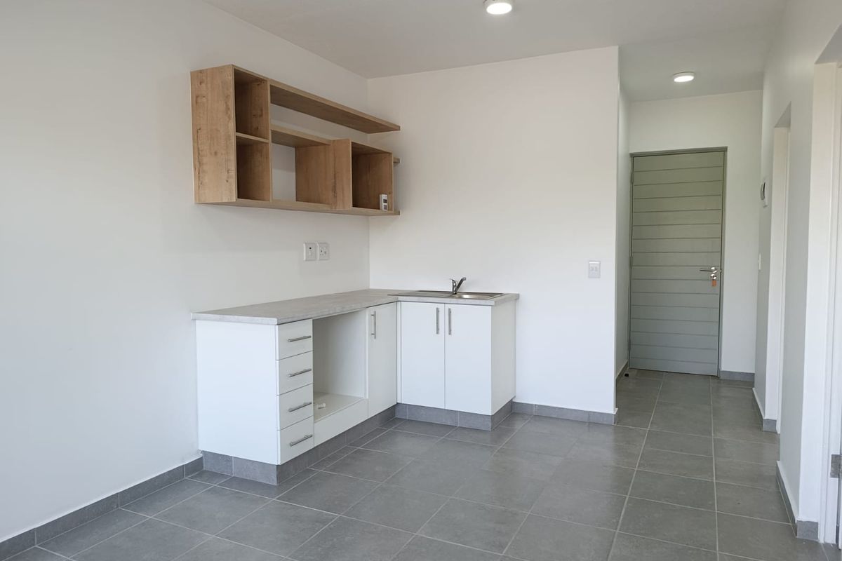 A clean, modern kitchen area featuring white cabinetry, wood-look shelving, grey tiled flooring, and a stainless steel sink, presented in a vacant, well-maintained state.
