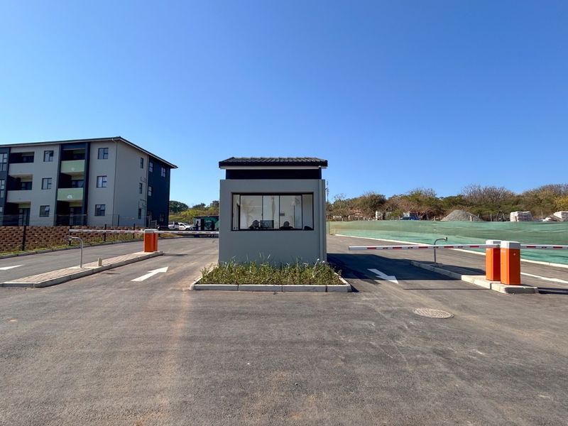 A modern apartment complex entrance featuring a security guard house, automated boom gates, and paved access roads.