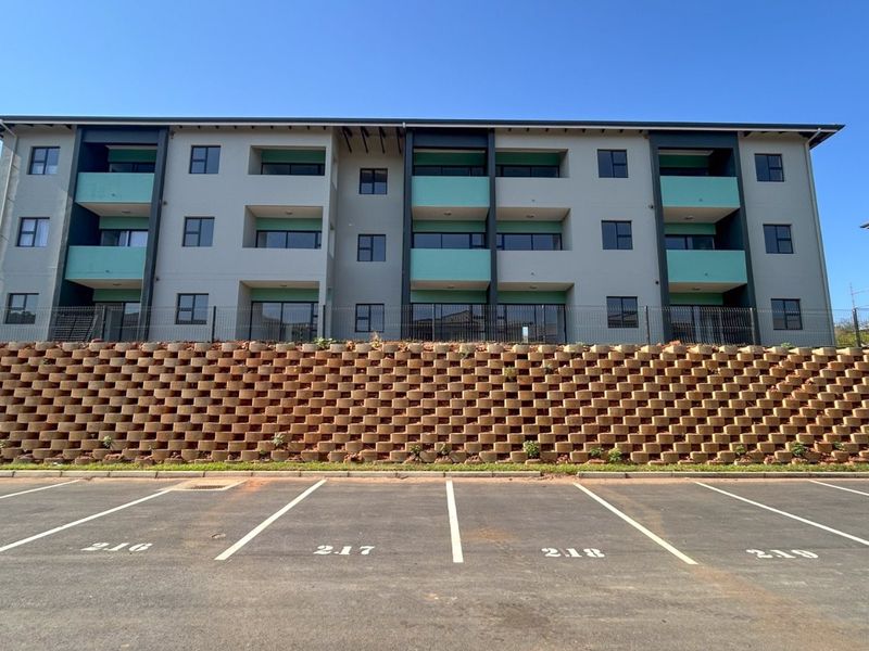Modern apartment building exterior featuring a large tiered retaining wall, designated asphalt parking bays, and private balconies with sliding door access.