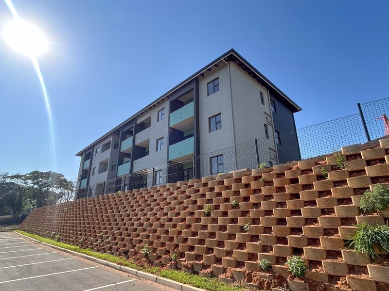 Exterior view of a modern apartment building featuring balconies, secure perimeter fencing, and a large tiered retaining wall overlooking a paved parking area.