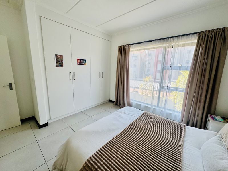 A bright bedroom featuring neutral tiled flooring, ample built-in cupboard storage, and a large window allowing for natural light.