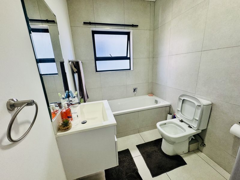 A modern, well-maintained bathroom featuring neutral floor-to-ceiling tiling, a built-in bathtub, a wall-mounted vanity, and a contemporary toilet.